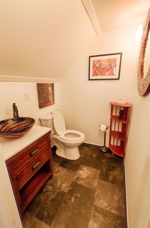Un baño con tocador de madera, inodoro blanco, un cuadro en la pared y una pequeña estantería con un jarrón.