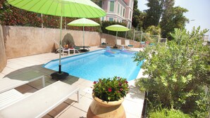 Une piscine entourée de chaises longues et de parasols, elle-même bordée d’un jardin et dominée, au loin, par un grand bâtiment.