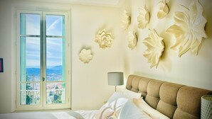 Une chambre dotée d’une grande fenêtre, d’un lit recouvert de draps blancs, d’une tête de lit beige en tissu et de décorations florales fixées au mur.