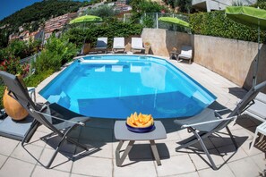 Une piscine entourée de chaises longues et de parasols, avec une végétation luxuriante et des bâtiments en arrière-plan.