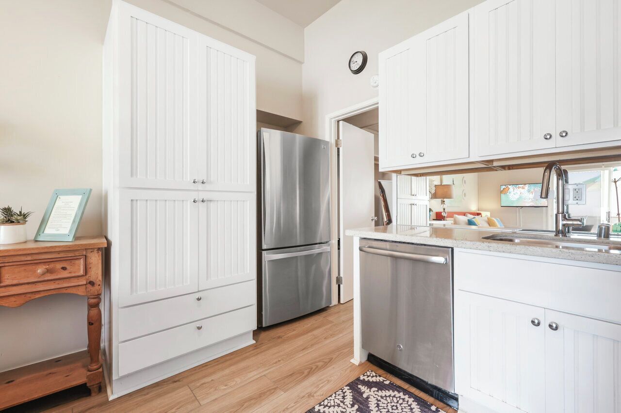 A kitchen with white cabinetry, a stainless steel refrigerator, a dishwasher, and a wooden table with a plant and a framed certificate.