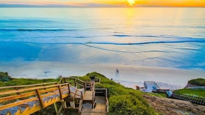 A coastal view with a wooden staircase leading to a beach, a clear sky, and the sun setting over the water.