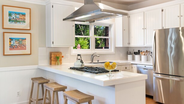 A modern kitchen with white cabinetry, a stainless steel range hood, a built-in island with stools, and a large refrigerator. - Charming Farm House Downtown St. Helena!  3bedrooms/3baths
Permit # STR26-014 (St Helena)