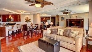 A spacious living area with a sofa, coffee table, and a dining table with chairs. There is a kitchen area with wooden cabinets and a ceiling fan. - Premier Ocean View! King Luxury Bed, A/C, Clean, Cozy, Updated, Wifi, Beach Gear (Kihei)
