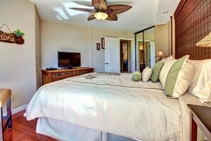 A bedroom with a bed, a television on a wooden cabinet, a ceiling fan, and a door leading to another room. - Premier Ocean View! King Luxury Bed, A/C, Clean, Cozy, Updated, Wifi, Beach Gear (Kihei)