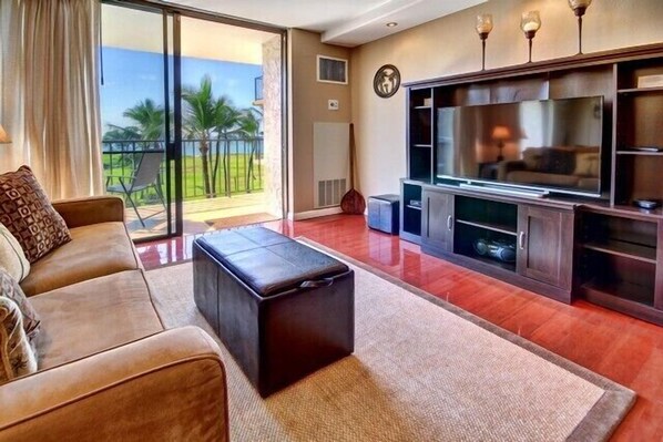 A living room with a sofa, a coffee table, a flat-screen TV, and a sliding glass door leading to a balcony with a view of palm trees. - Premier Ocean View! King Luxury Bed, A/C, Clean, Cozy, Updated, Wifi, Beach Gear (Kihei)