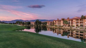 Un parcours de golf avec un étang dans lequel se reflètent les bâtiments, le tout étant entouré de montagnes.