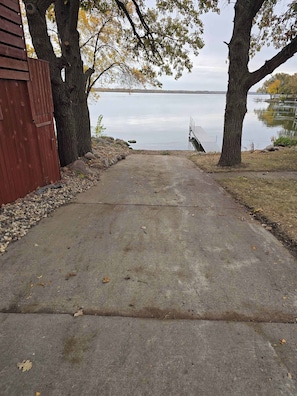 A concrete pathway leading to a body of water, flanked by trees and a red building.