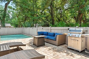 A backyard with a pool, a blue outdoor sofa, a grill, and a dining area with chairs and a table.