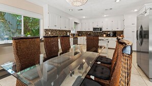 Una cocina moderna con gabinetes blancos, una mesa de comedor de vidrio, sillas de mimbre y un refrigerador de acero inoxidable.