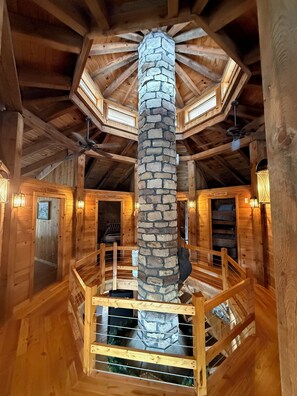 A wooden cabin interior with a stone fireplace, wooden beams, and a spiral staircase.