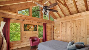 Ein Schlafzimmer einer Holzhütte mit Bett, Deckenventilator, einem roten Sessel und großen Fenstern.