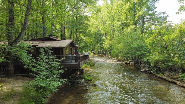 A cabin with a deck overlooking a river, surrounded by dense forest. - River's Edge - Ultimate Riverfront Cabin w/Hot Tub (Helen)