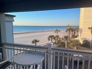 A balcony with a view of a beach, palm trees, and a parked car. - Oceanfront Condo • 2026 Renovations! • 2 BD, 2BA (Myrtle Beach)
