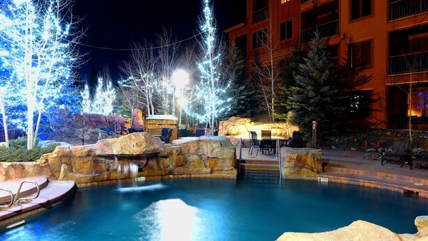 A nighttime outdoor pool area with a waterfall, surrounded by rock formations and trees adorned with lights. - Family Friendly in River Run. Heated pool & slide, steps from gondola and shops (Keystone)