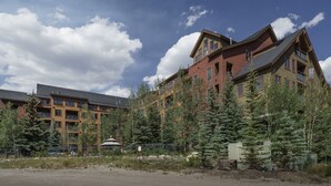 A multi-story building with a mix of red and brown exterior walls, surrounded by evergreen trees. - Family Friendly in River Run. Heated pool & slide, steps from gondola and shops (Keystone)