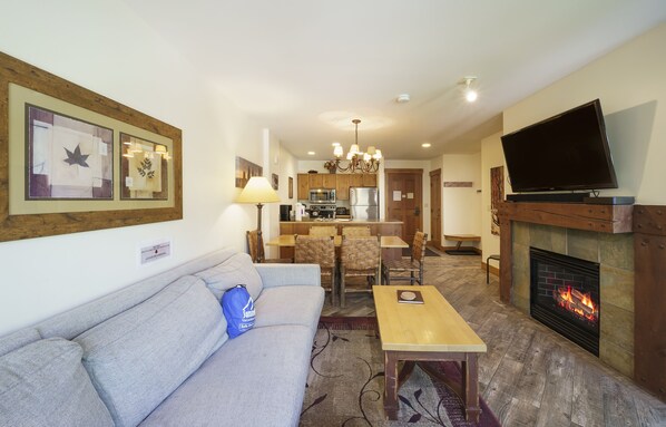 Een woonkamer met een bank, een salontafel en een open haard. Er is een televisie aan de muur bevestigd en een keuken in de achtergrond.