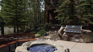 A stone fire pit with a metal grill, surrounded by a stone wall and a wooden fence. - Family Friendly in River Run. Heated pool & slide, steps from gondola and shops (Keystone)