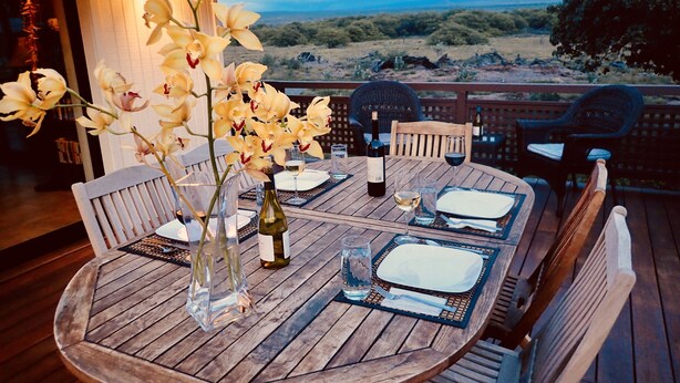 Une terrasse en bois avec une table ronde dressée pour le dîner, agrémentée d’assiettes blanches, de verres à vin et d’une bouteille de vin.