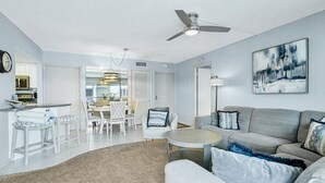 Un salon moderne avec un canapé gris, une table basse ronde et un coin repas comprenant des chaises blanches et un lustre.