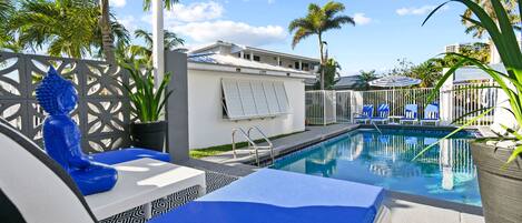 Zona de piscina con sillones de descanso, una palmera y un edificio al fondo.