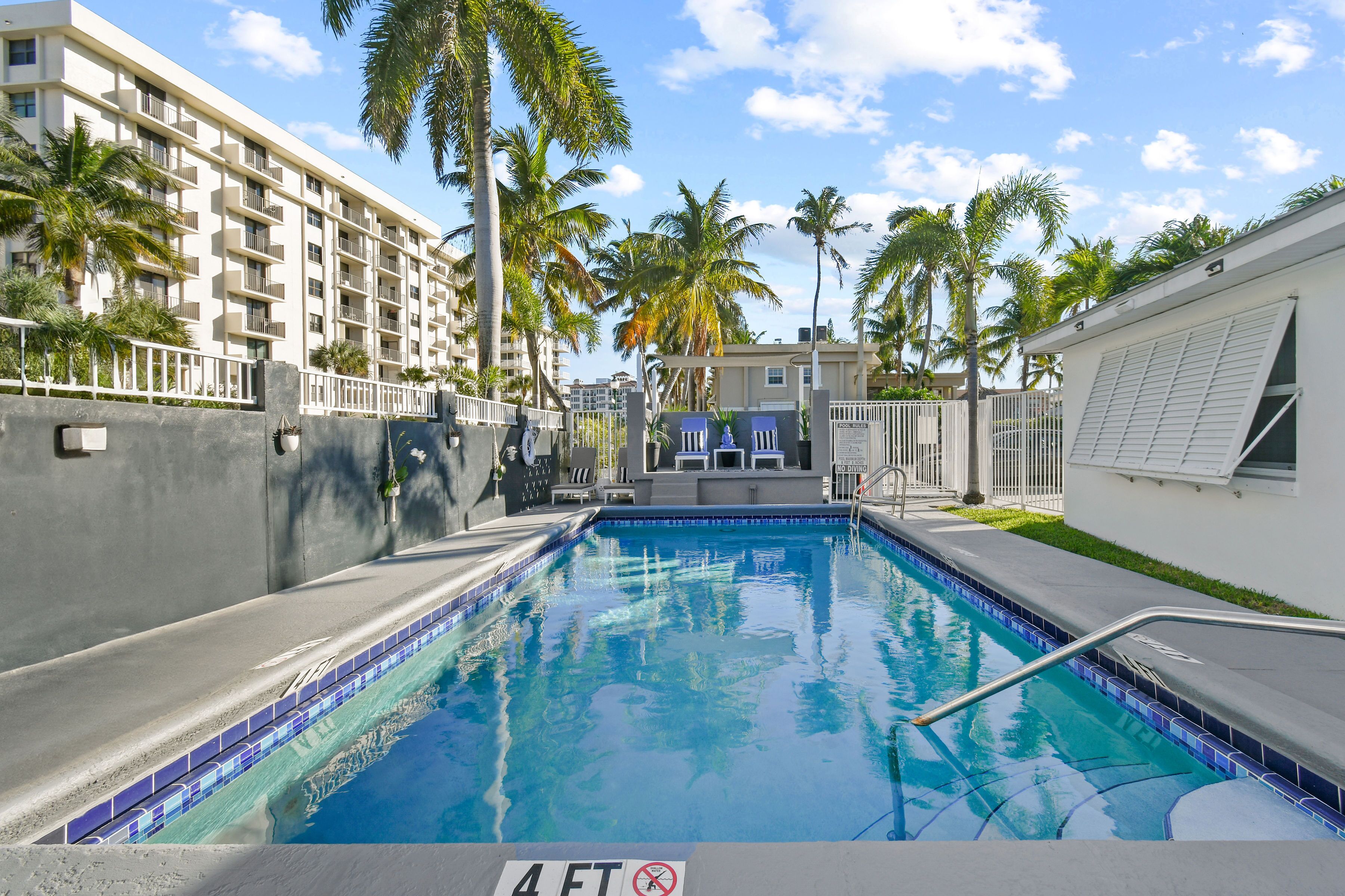 A swimming pool with a 4 FT depth limit, surrounded by palm trees and a multi-story building.