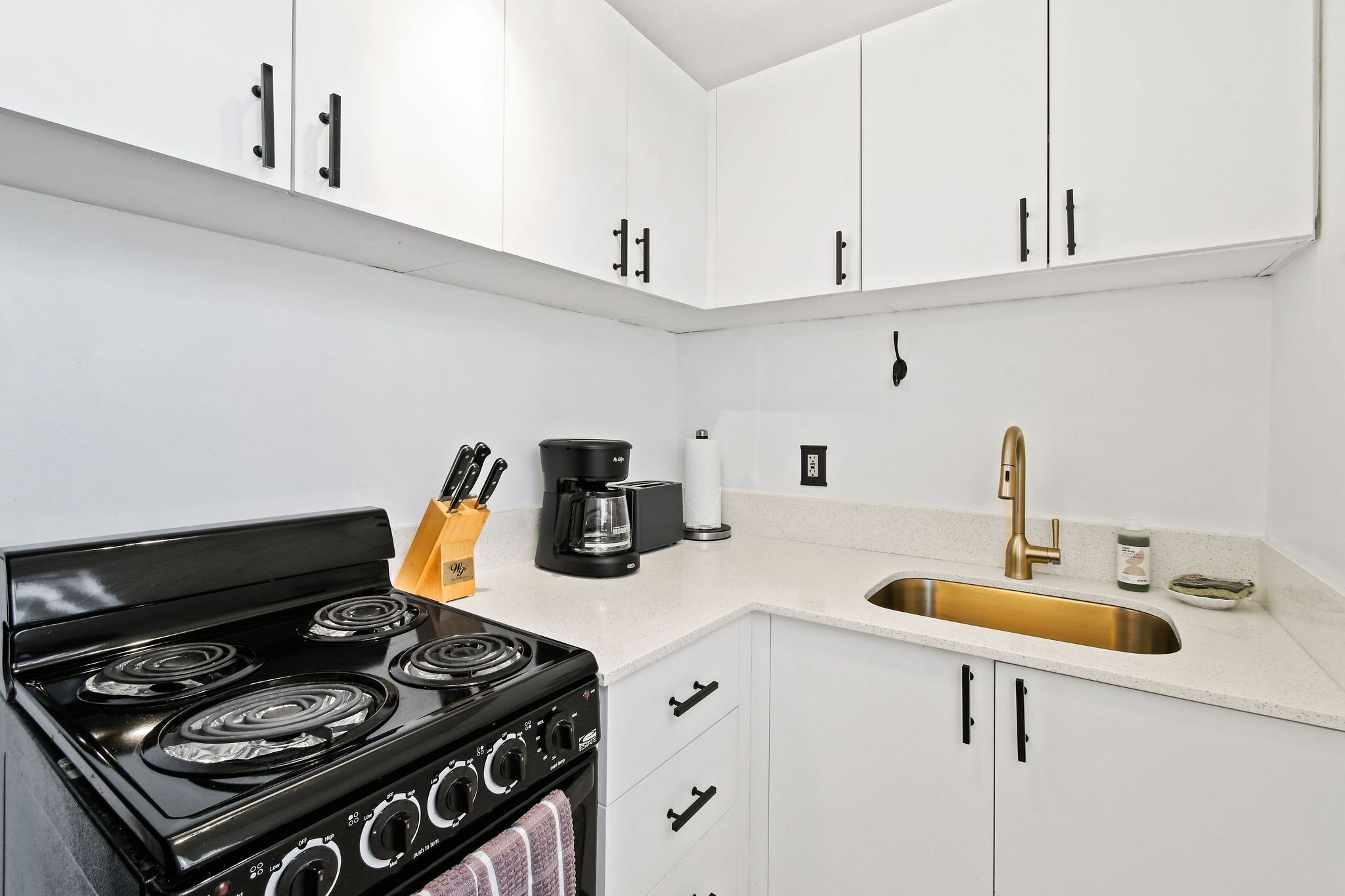 A modern kitchen with white cabinets, a black stove, a coffee maker, and a gold faucet.