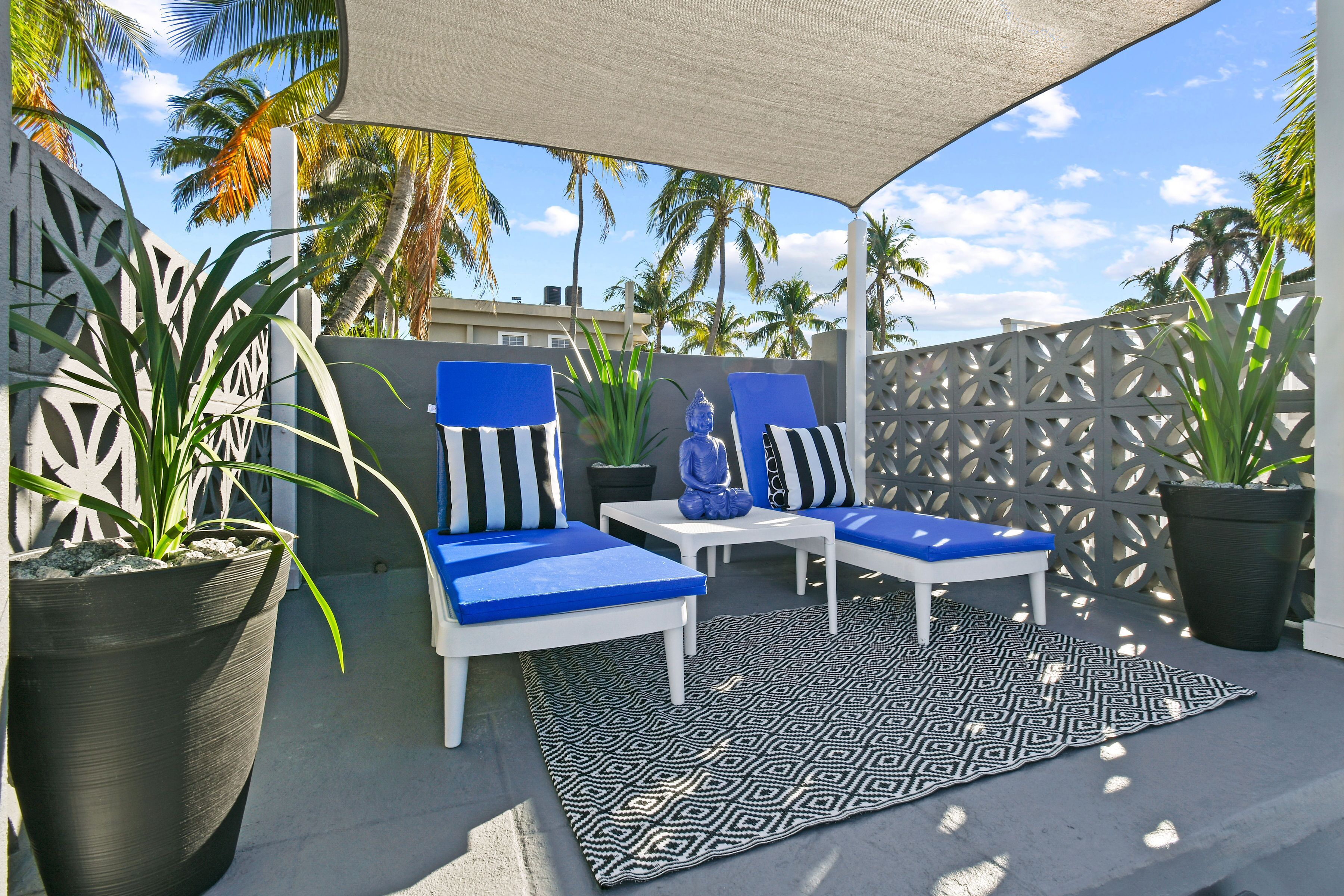 A covered outdoor seating area with two chairs and a table, surrounded by potted plants and palm trees.