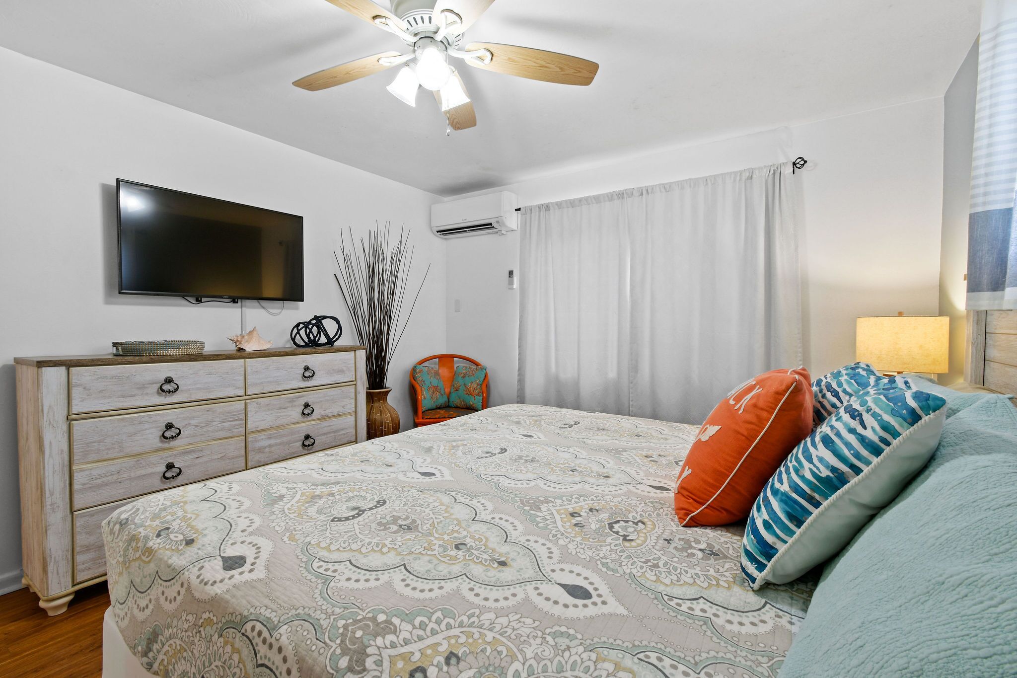 Une chambre à coucher comprenant un lit, une commode, un téléviseur, un climatiseur, un ventilateur de plafond et une fenêtre avec des rideaux.