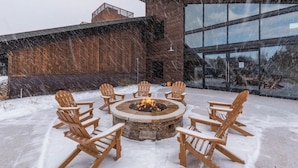 Ein runder Feuerkorb mit sechs Adirondack-Stühlen darum herum, auf einer verschneiten Terrasse, im Hintergrund ein modernes Gebäude.