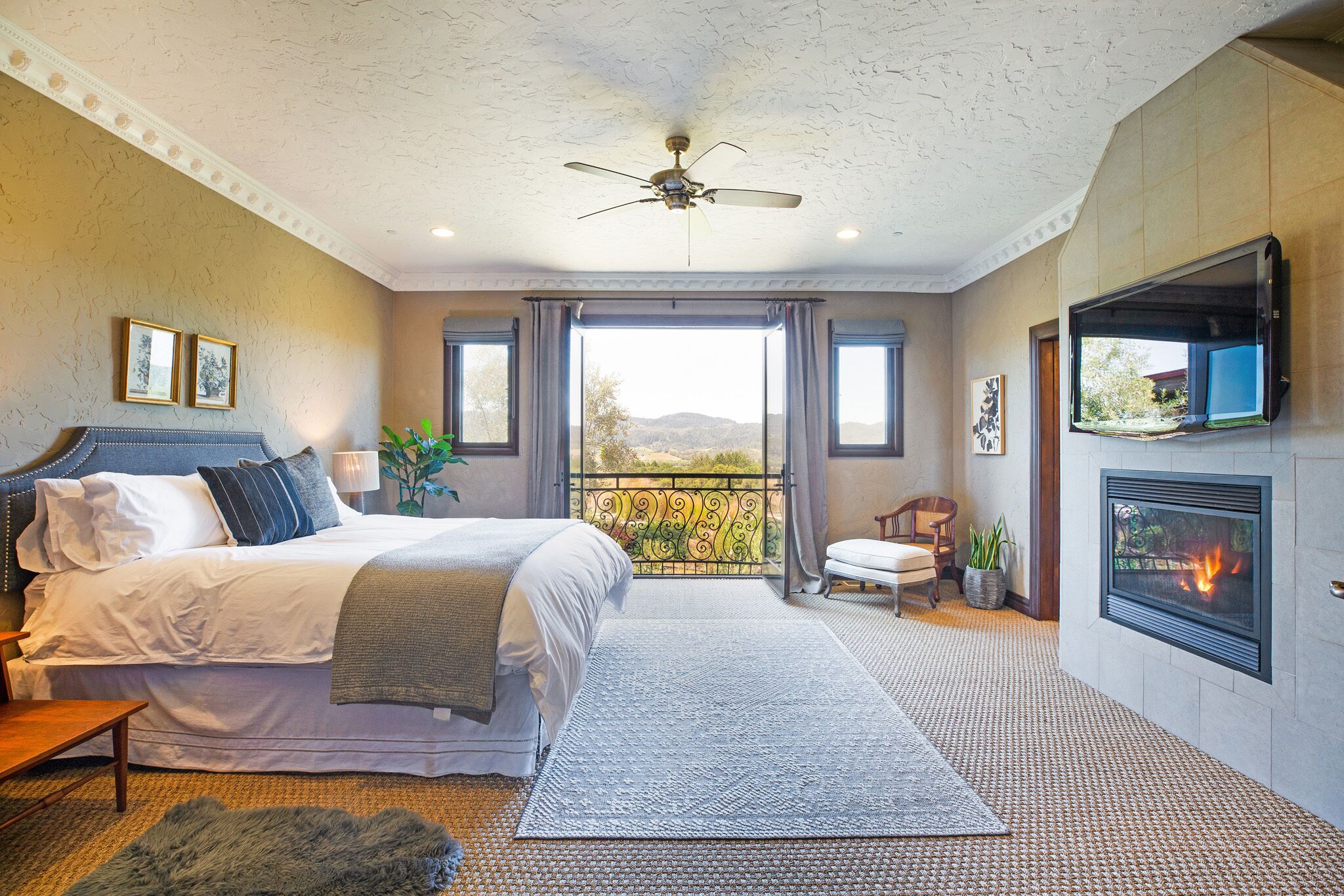 A bedroom with a bed, a ceiling fan, a fireplace, and a view of the outdoors.