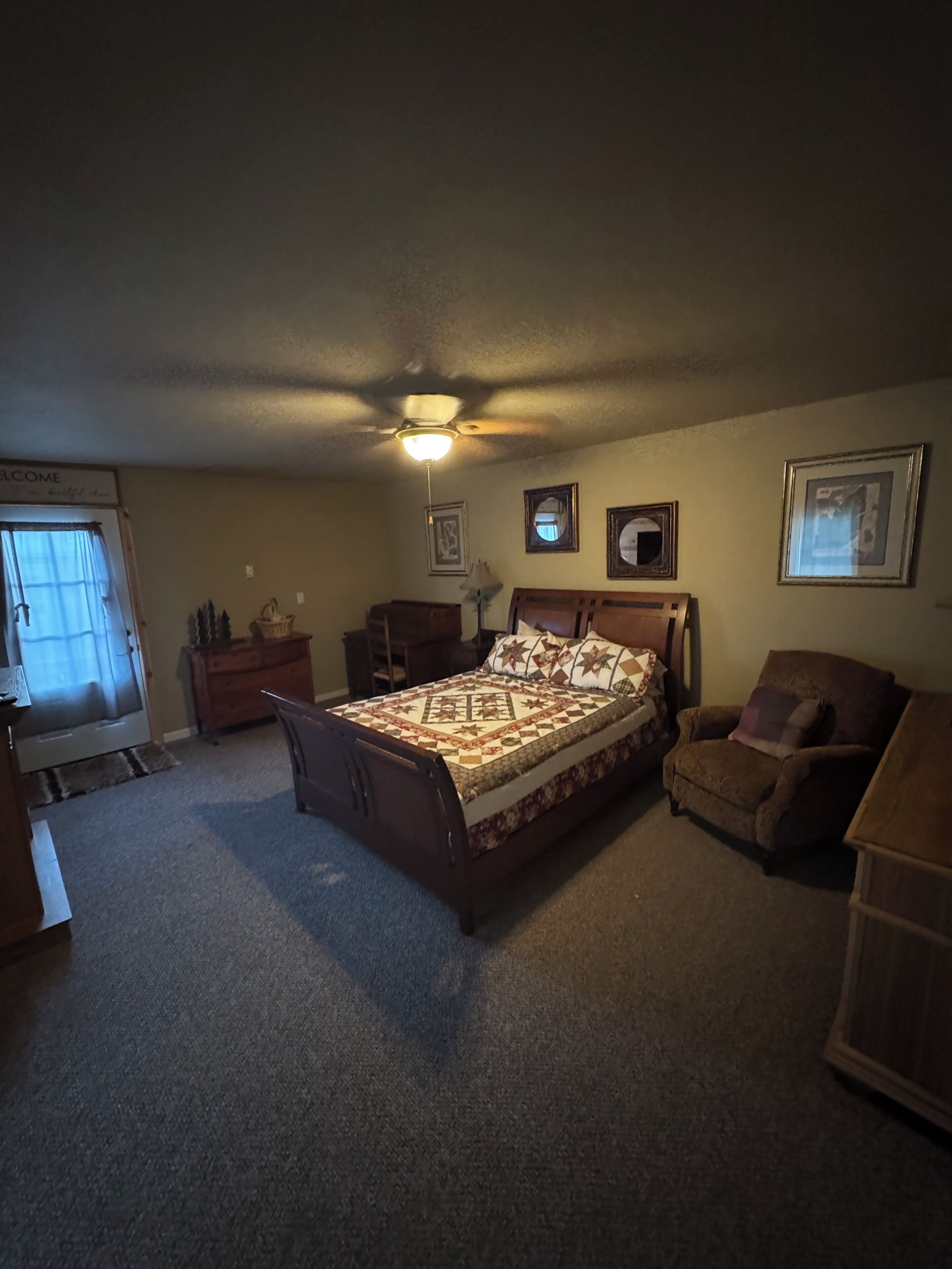 A bedroom with a bed, a chair, a desk, a ceiling fan, and framed pictures on the wall.