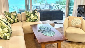 Ein Wohnzimmer mit einer Couch, Sesseln und einem Couchtisch. Durch große Fenster ist der Blick auf das Meer und Palmen.