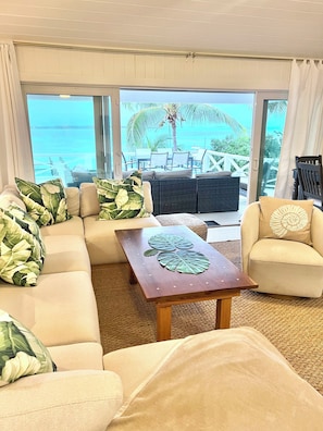 A living room with a sofa, armchairs, and a coffee table. There is a view of the ocean and palm trees through large windows.