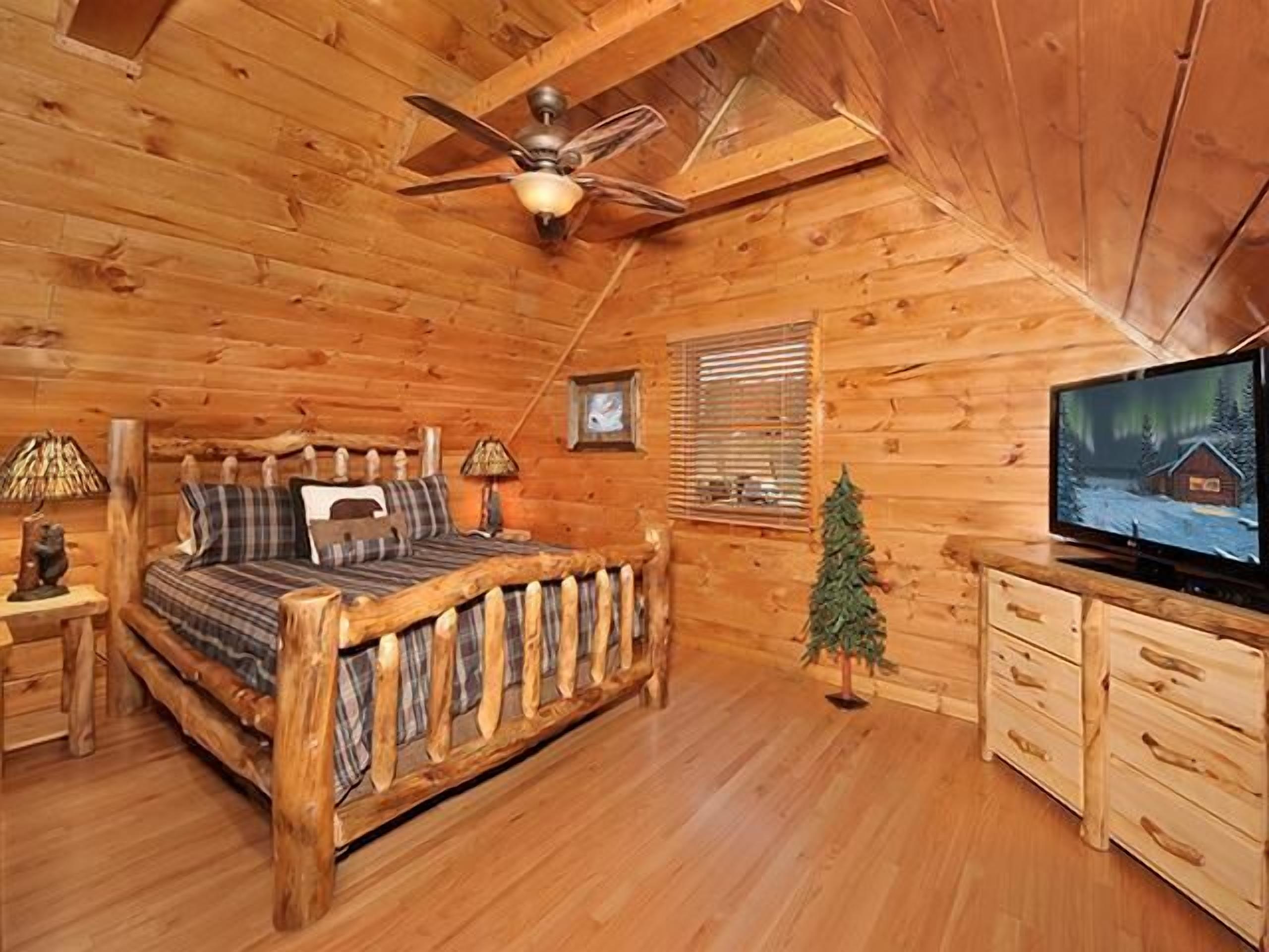 Une chambre dans un chalet en rondins, avec un lit en bois, un ventilateur de plafond, une télévision, un petit sapin de Noël et une fenêtre avec des stores.