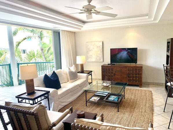 A living room with a sofa, coffee table, TV, and a balcony with a view of palm trees. - The Best of Everything in Providenciales, Part II (Providenciales)