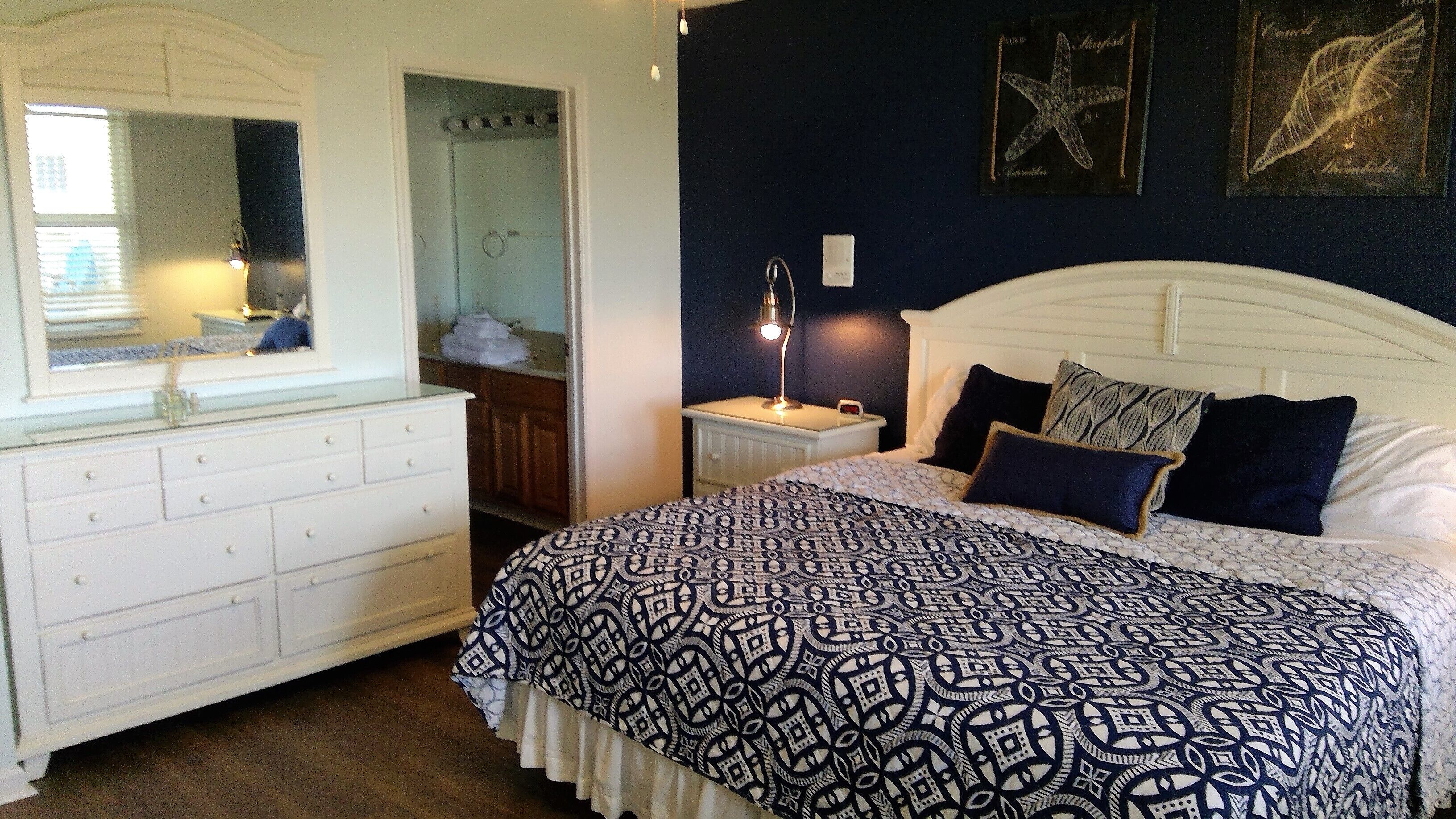 A bedroom with a white dresser, a bed with patterned bedding, a nightstand with a lamp, and a dark blue wall with framed artwork.
