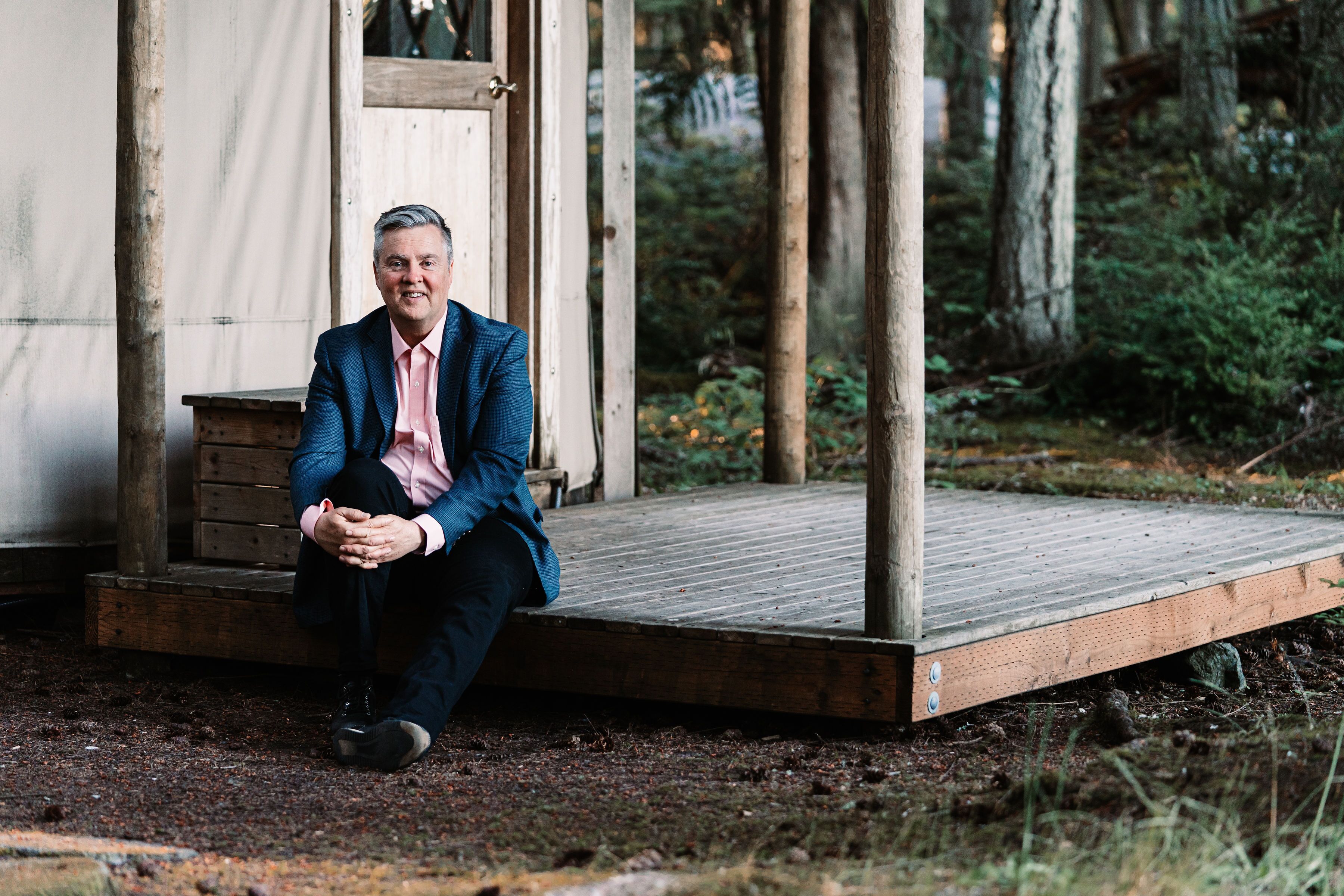 A man sitting on a wooden platform outside a rustic building surrounded by trees.