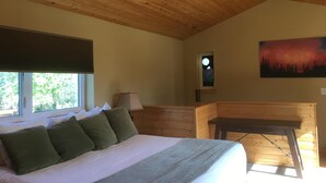 A bedroom with a wooden ceiling, a bed with green pillows, a nightstand, a lamp, a window with a view of trees, and a painting on the wall.