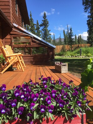 Una terraza de madera con una silla de madera, una jardinera con flores moradas y vistas a una zona boscosa.