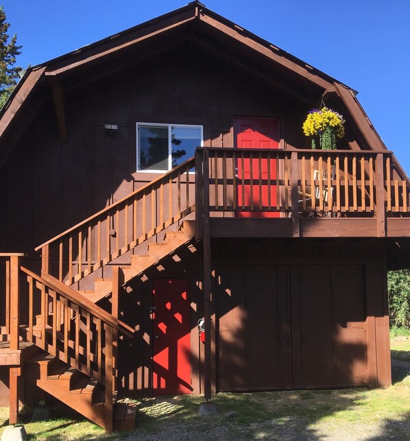 Un edificio de estilo granero de madera con puerta roja, un balcón pequeño con arreglo floral y una escalera que conduce a la entrada.