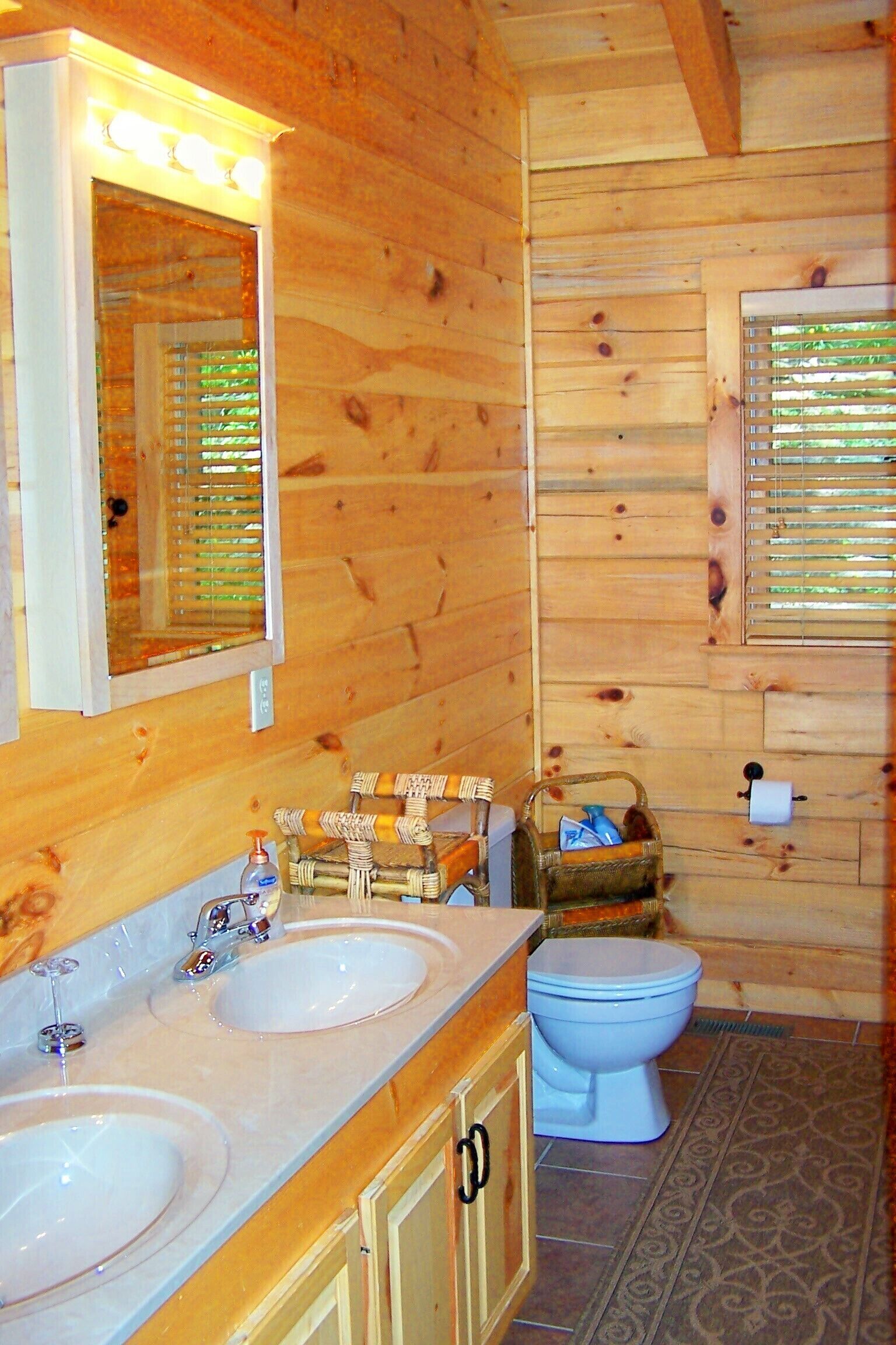 A wooden cabin bathroom with two sinks, a mirror, a toilet, and a window with blinds.