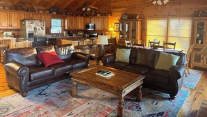 A cozy cabin-style interior with a wooden ceiling, a dining area, a kitchen, and a living room with leather sofas and a coffee table.