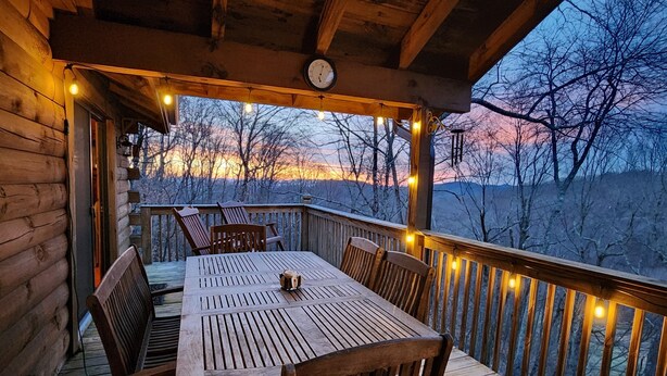 A wooden deck with a table and chairs, string lights, and a view of trees and hills.