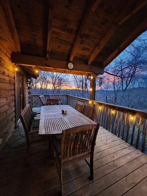 Une terrasse en bois avec une table et des chaises, des guirlandes lumineuses et une vue sur les arbres et les collines.