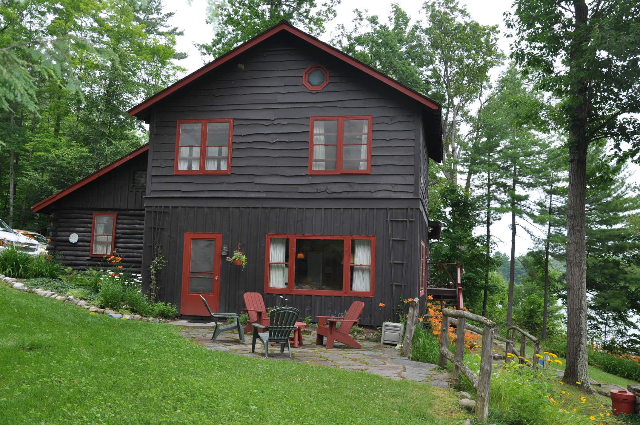 Une cabane en rondins avec un toit rouge et deux fenêtres, entourée d’un jardin agrémenté de chaises et d’une table.