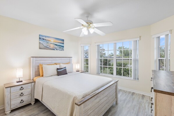 A bedroom with a large bed, a ceiling fan, a nightstand with a lamp, and a window with a view of greenery.