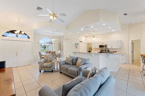 A spacious living area with a sofa, armchairs, and a dining table. A kitchen with white cabinets and a central island is visible in the background.