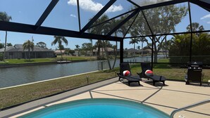 Zona de piscina con sillones reclinables y una parrilla, con vistas a una masa de agua y una zona residencial.