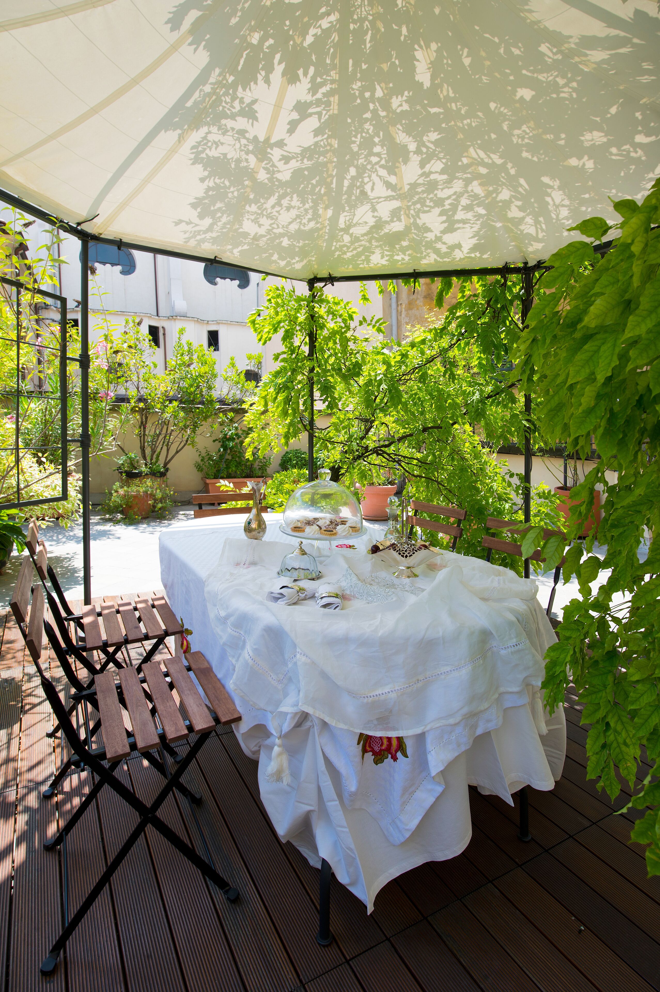 A covered patio with a table set for a meal, surrounded by greenery and potted plants.
